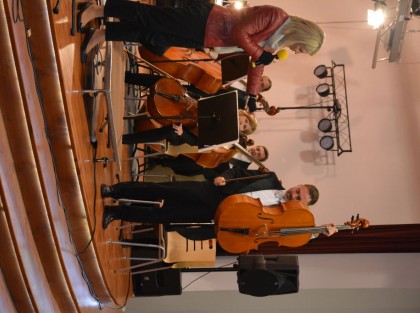 kobieta w czerwonej kurtce a obok muzyk pokazujący wiolonczelę